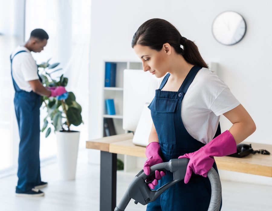 Selective focus of attractive worker of cleaning service using vacuum cleaner near african american